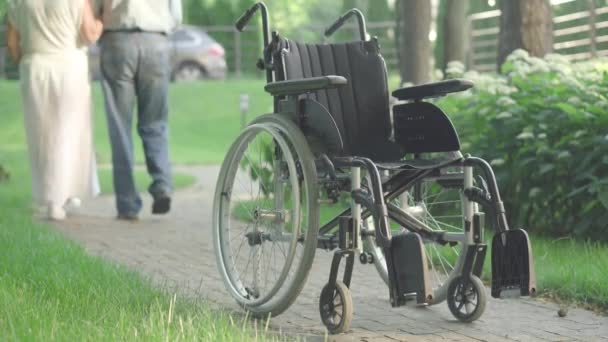 Rolstoel die in het zonlicht op een steegje in het park staat met wazig ouder blank echtpaar dat op de achtergrond wegloopt. Oude man en vrouw wandelen buiten met zwakke echtgenoot op zomerdag. — Stockvideo