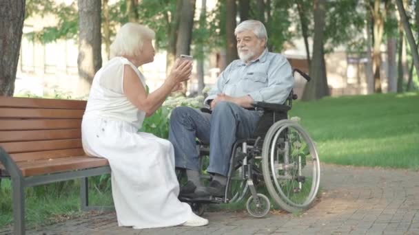 Wide shot of positive smiling husband and wife using smartphone in sunny park. Loving senior Caucasian woman sitting on bench with disabled husband in wheelchair and talking. — Stock Video