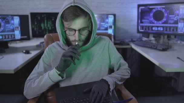 Confident man in eyeglasses and black gloves smoking and looking at camera. Portrait of concentrated Caucasian hacker using laptop and multiple computers in office at night for stealing information. — Stock Video