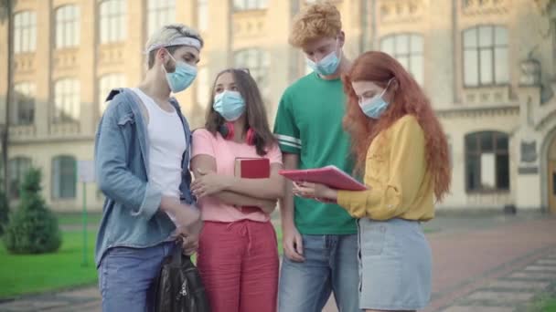Fyra universitetsstudenter i ansiktsmasker diskuterar läxprojekt utomhus. Grupp unga kaukasiska män och kvinnor som står på campus och pratar under Covid-19-pandemin. Utbildning i coronavirus. — Stockvideo