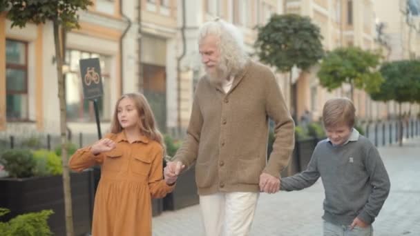 Portrait of senior man walking with grandchildren and listening to excited kids talking. Old grey-haired Caucasian grandfather enjoying summer weekend with cute happy boy and girl outdoors. — Stock Video