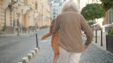Kafkasyalı büyükbaba ve torununun şehir caddesinde gezinip dans edişinin arka görüntüsü. Sevimli şirin kız, sevimli büyükannenle büyükbabana sarılıyor ve gülümsüyor. Mutluluk ve çocukluk.