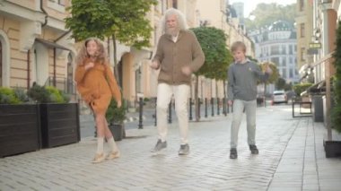 Güneşli şehir caddesinde neşeli büyükbaba ve torunların komik dansı. Neşeli son sınıf öğrencisinin geniş açılı portresi, açık havada dans eden küçük oğlan ve kız. Eğlence ve eğlence konsepti.