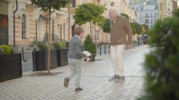 Glad liten pojke som springer med fotbollsboll till leende senior man. Glädjefyllda vita barnbarn och farfar på stadens gata. Glada barn angelägna om sport njuter helger med mor- och farförälder utomhus. — Stockvideo