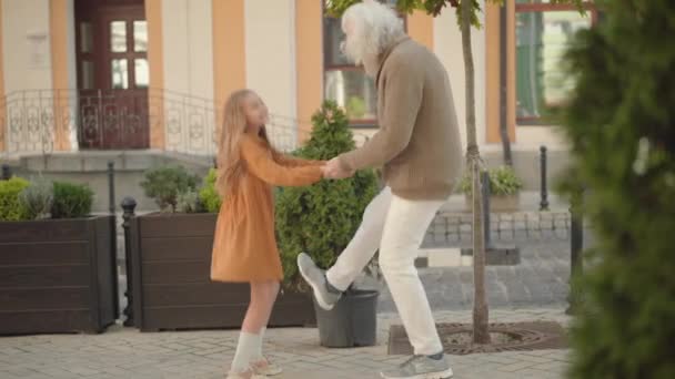 在城市街道上与年长的快乐男人共舞的快乐可爱女孩的全景。一个快乐的白人孙女和祖父的画像，在阳光明媚的户外享乐。快乐与快乐. — 图库视频影像