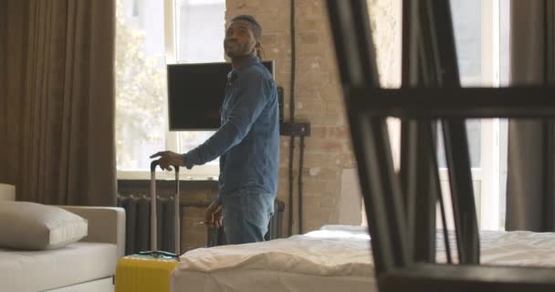 Radostně odpočatý Afroameričan se zavazadly rozhlížející se po hotelovém pokoji a padající zpátky na postel. Pozitivně se usmívající pohledný mužský turista těší dovolenou. Cestovní ruch a cestování. — Stock video