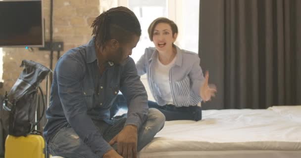 Jóképű afro-amerikai férfi ül az ágyon dühös kaukázusi nővel, gesztikulál és kiabál a háttérben. Boldogtalan barát és barátnő, akiknek kapcsolati nehézségeik vannak utazás közben. — Stock videók