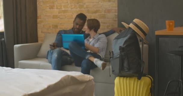 Packat bagage står i sovrum eller hotellrum med suddig interracial par surfa Internet på laptop i bakgrunden. Glad ung man och kvinna kontrollerar flygtid online. — Stockvideo