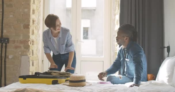 Positivo jovem casal bagagem de embalagem em saco de viagem amarelo no quarto. Mulher bonita branca sorridente colocando chapéu de palha e homem afro-americano bonito mostrando o polegar para cima. Viagens e turismo — Vídeo de Stock