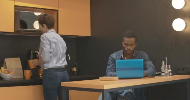 Occupé copain afro-américain travaillant en ligne sur ordinateur portable comme petite amie caucasienne faire du thé sur la cuisine. Heureux couple interracial détendu de pigistes à la maison. — Video