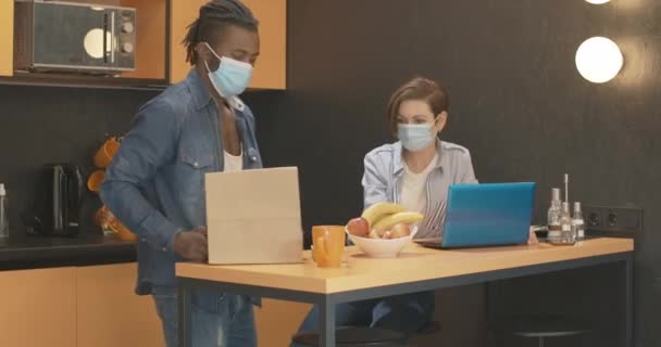 Межрасовая пара в маске Ковид-19 разговаривает дома на кухне. Уверенная белая женщина и афроамериканец вечером в помещении. Пандемический образ жизни коронавируса. — стоковое видео
