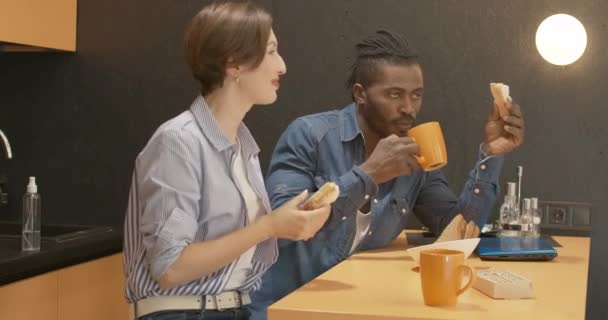 Magabiztos afro-amerikai férfi portréja, amint szendvicset eszik és teát iszik, miközben kaukázusi nő beszél elöl. Fiatal boldog fajok közötti pár reggelizik a konyhában. — Stock videók