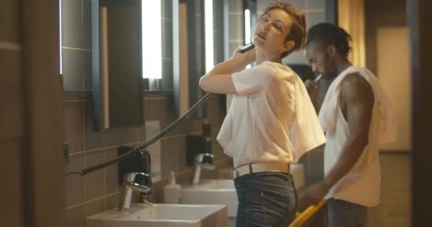 Młoda kobieta suszenie włosów z suszarką jak mężczyzna mycie zębów w tle. Widok z boku kaukaska dziewczyna i Afroamerykanin chłopak w łazience rano. — Wideo stockowe