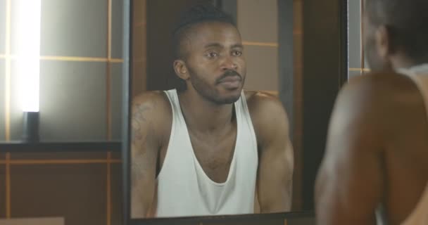 Reflet d'un jeune Afro-Américain regardant dans un miroir et réfléchissant. Beau garçon réfléchi avec tatouage à portée de main quittant la salle de bain le matin. — Video