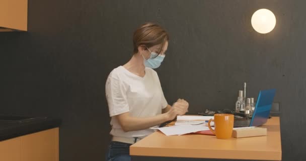 Covid-19フェイスマスクでノートパソコンの自撮りカメラに手を振って書き込み中の若い女の側面図。コロナウイルスのパンデミック発生時にオンラインで働くブルネットの女性従業員. — ストック動画