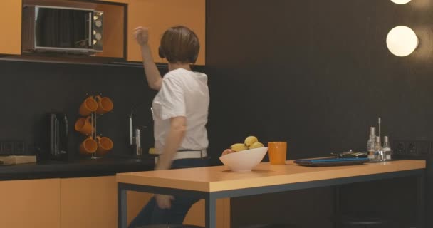 Joven mujer alegre en auriculares bailando en la cocina y saliendo con manzana. Retrato de dama caucásica alegre y despreocupada disfrutando de una noche feliz en casa comiendo frutas saludables y escuchando música. — Vídeo de stock