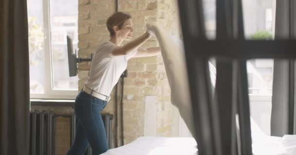 Joven mujer alegre haciendo la cama por la mañana y cayendo de nuevo en manta blanca. Alegre señora caucásica sin preocupaciones disfrutando de fines de semana o vacaciones en el dormitorio en casa. — Vídeos de Stock