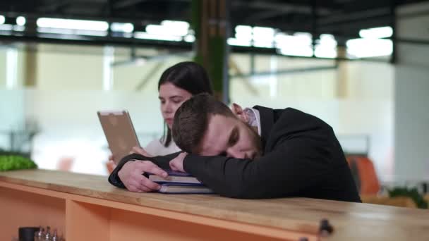 Empresário exausto dormindo na mesa com empresária turva navegando na Internet em tablet ao fundo. Cansado jovem caucasiano acordando e esfregando os olhos. Conceito de excesso de trabalho. — Vídeo de Stock