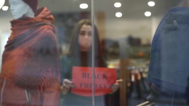 Jonge blanke vrouwen naderen de etalage en hangen aankondigingen op over zwarte vrijdagverkoop. Portret van twee winkelmedewerkers in Covid gezichtsmaskers die kleding verkopen tijdens een coronaviruspandemie. — Stockvideo