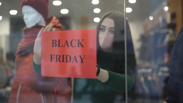 Junge brünette Frau mit Covid-19-Gesichtsmaske, die die Verkaufsankündigung für den Black Friday am Schaufenster aufhängt. Porträt einer konzentrierten Frau, die während der weltweiten Virenpandemie in einem Einkaufszentrum arbeitet. — Stockvideo