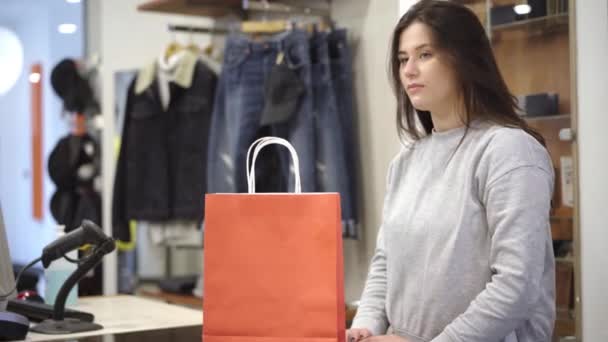 Jonge mooie blanke vrouw passeert boodschappentassen bij de kassa. Positieve brunette winkelassistent werkt in de winkel. Concept zakelijke dienstverlening en consumentisme. — Stockvideo
