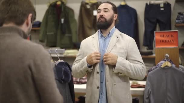 Przystojny brodaty biały mężczyzna przymierza kurtkę w sklepie i przyjaciel aprobuje swój wybór. Pewny siebie poważny mężczyźni wybierając ubrania w centrum handlowym podczas sprzedaży czarny piątek. — Wideo stockowe