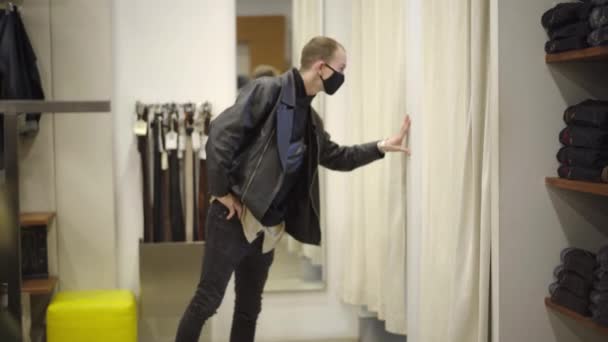 Портрет молодого чоловіка в Ковид-19, який чекає на друзів, які намагаються одягнути одяг в гардеробній кімнаті. Хлопець, що показує пальці на колесах в новому спорядженні. Концепція купівлі Коронавірусу. — стокове відео