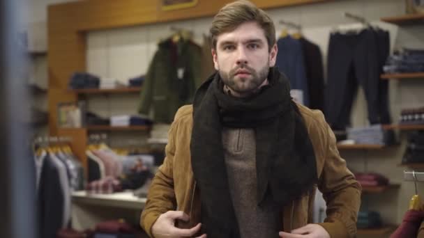 Guapo joven con estilo probándose la ropa en la tienda. Retrato de hombre caucásico comprador en chaqueta y bufanda mirando a la cámara como espejo y sonriente. Concepto de confianza y compras. — Vídeo de stock