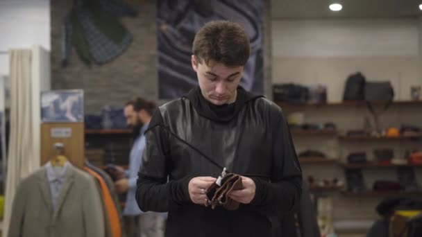 Joven hombre caucásico mirando la billetera de cuero caro y salir de la tienda. Retrato de comprador masculino insatisfecho eligiendo mercancías en el centro comercial. Compra y comercio. — Vídeo de stock