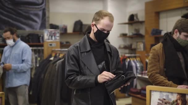 Młody mężczyzna w masce Covid-19 kichający w sklepie odzieżowym dla mężczyzn, klienci uciekający przed zakażonym klientem. Problemy z zakupem koronawirusów podczas globalnej pandemii wirusowej. Koncepcja strachu i paniki. — Wideo stockowe