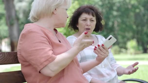 Vista lateral de una mujer rubia moderna jubilada mostrando las redes sociales a una amiga y morena arrogante haciendo un gesto con una expresión facial repugnante. Concepto de estilo de vida jubilatorio. — Vídeos de Stock