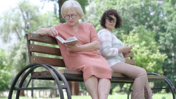 Porträtt av bekymmerslös vit senior kvinna läser bok som vän med smartphone kikar från bakgrunden. avslappnad intelligent dam tittar tillbaka på pensionär surfa Internet och suckande. Livsstil. — Stockvideo
