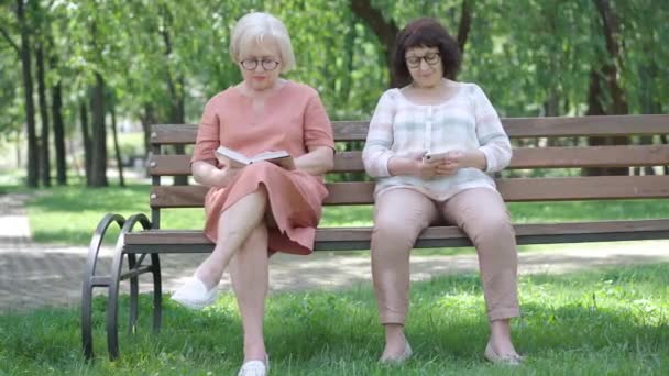 Breed portret van twee oudere vrouwen met hobby 's op de bank in het zonnige park. Geabsorbeerde blanke gepensioneerden lezen boek en surfen sociale media op zomerse dag buiten. Levensstijl concept. — Stockvideo