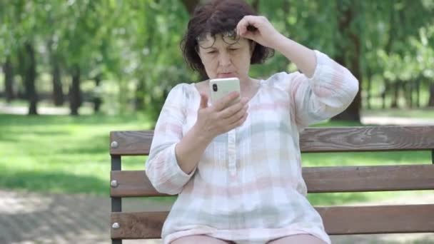 Mujer mayor con mala vista mirando la pantalla del teléfono inteligente en Sunny Park. Retrato de mujer caucásica jubilada en gafas de mensajería en línea. Concepto médico y de envejecimiento. — Vídeos de Stock