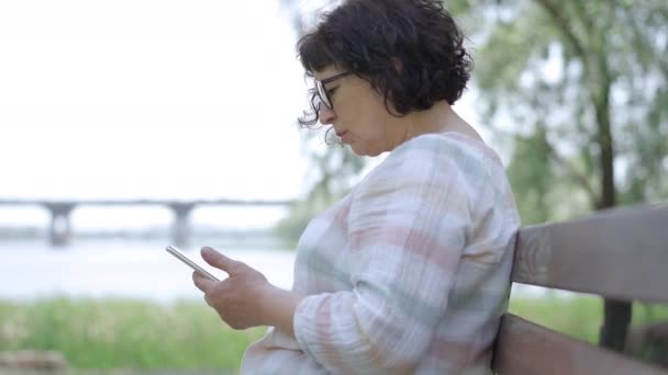 Vista lateral de la mujer mayor caucásica en gafas que se desplaza por las redes sociales en el teléfono inteligente. Moderno jubilado caucásico navegando por Internet. Mensajería de dama miope en línea. — Vídeo de stock