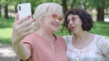 Güneş gözlüklü, güneşli yaz parkında selfie çeken kaygısız Kafkas emeklilerine yakın çekim. Gülümseyen ve dışarıda fotoğraf çeken neşeli, rahat kadınların portresi. Mutlu emeklilik kavramı.