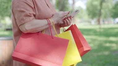 Alışveriş çantalarıyla akıllı telefondan sosyal medyaya kayan tanınmayan emeklilerin yan görüntüsü. Rahat, modern Kafkas kıdemli kadın açık havada güneşli yaz parkında internetten mesajlaşıyor..