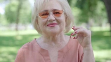 Gülümseyen beyaz bir kadının yakın plan portresi. Güneş gözlüğü takıyor ve kameraya göz kırpıyor. Güneşli yaz parkında neşeli bir emeklinin flört etmesi. Neşe ve yaşam tarzı kavramı.