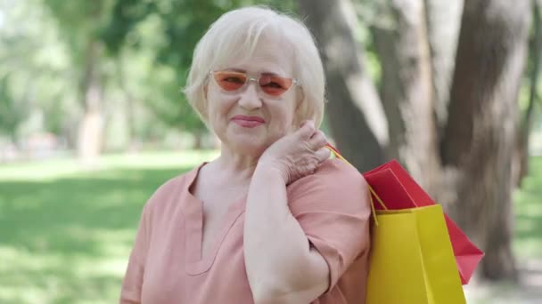 Retrato de jubilado caucásico feliz posando con bolsas de compras al aire libre. Mujer mayor alegre en gafas de sol mirando a la cámara y sonriendo. Elegante concepto de envejecimiento y jubilación. — Vídeos de Stock