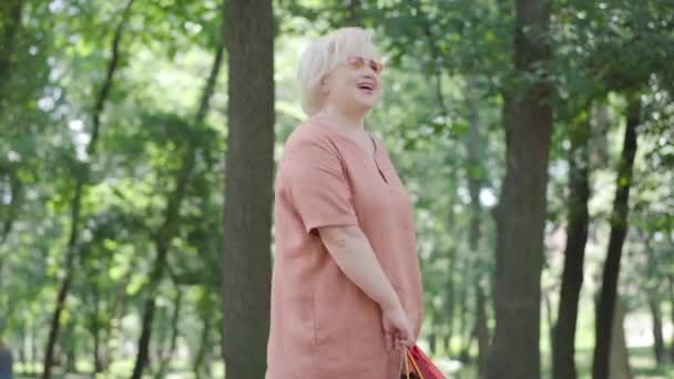 快乐的资深白人退休人员带着购物袋在阳光灿烂的夏季公园里旋转。一副戴着太阳镜的快乐无忧无虑的漂亮女人的画像，令人喜形于色。1.购物主义和生活方式概念. — 图库视频影像