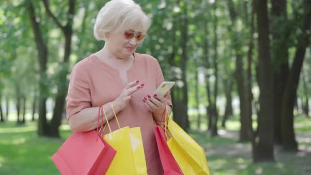 Elegante mujer mayor con bolsas de compras de mensajería en línea utilizando el teléfono inteligente. Retrato de confianza hermosa jubilada caucásica en gafas de sol navegar por Internet en el parque de verano. Concepto de ocio. — Vídeo de stock