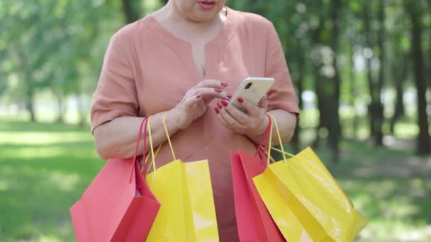 Onherkenbare senior vrouw met boodschappentassen surfen op internet op smartphone en praten. Happy Caucasian gepensioneerde berichten online en glimlachend. Geluk en vrije tijd concept. — Stockvideo