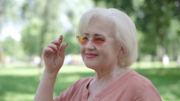 Gondtalan idős nő a kamera felé fordul, leteszi a napszemüveget, kacsint és mosolyog. Pozitív kaukázusi nyugdíjas portréja flörtöl a napos nyári napon a parkban. Boldogság és pihenés. — Stock videók