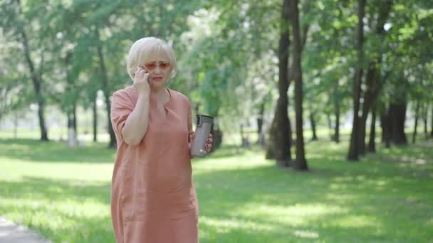 Retraite détendue marchant le long de l'allée dans un parc ensoleillé et parlant au téléphone. Portrait d'une femme blanche confiante et insouciante se promenant à l'extérieur avec un thermos à café. Concept de loisirs et de pension. — Video