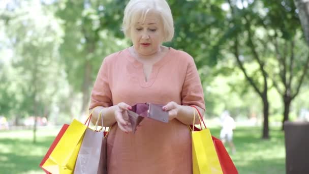 Starsza kobieta z torbami na zakupy patrzy na pusty portfel i wzdycha. Portret zdezorientowanego białego emeryta stojącego w słonecznym parku z zakupami i bez pieniędzy. Koncepcja zakupoholizmu. — Wideo stockowe