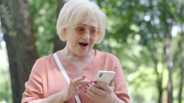 Podekscytowana starsza kobieta patrząca na ekran smartfona i uśmiechnięta. Portret niezwykle szczęśliwego białego emeryta w okularach przeciwsłonecznych wiwatującego na świeżym powietrzu w słoneczny letni dzień w parku. Koncepcja dobrych wiadomości. — Wideo stockowe