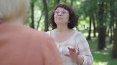 Güneş gözlüklü yaşlı esmer kadının, güneşli parkta sarışın arkadaşıyla sohbet edişinin portresi. Beyaz asıllı, emekli, yaz günü dışarıda kadın emeklilerle dedikodu yapıyor..