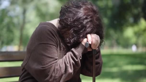 Huilende brunette senior vrouw zittend met wandelstok op bank in zomerpark. Depressieve blanke vrouwelijke gepensioneerde besteden eenzame pensionering dag buiten. Frustratie en eenzaamheid. — Stockvideo