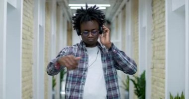 Ofis koridorunda dans eden gözlüklü ve kulaklıklı Afro-Amerikan genç adam. Kendine güvenen neşeli bir çalışanın iş yerinde eğlenen portresi. Sinema 4k ProRes Merkezi.