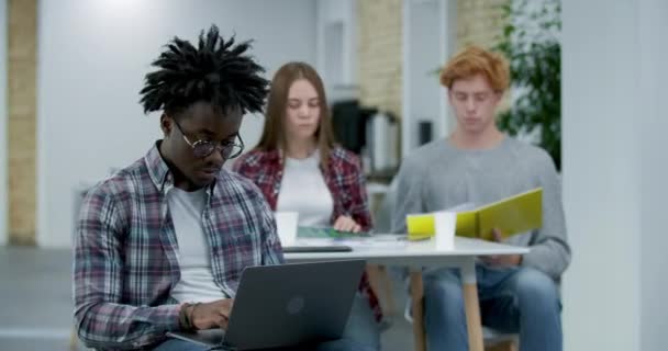 Travailleur de bureau afro-américain absorbé tapant sur le clavier de l'ordinateur portable, regardant la caméra et souriant. Portrait d'un homme positif concentré avec des collègues caucasiens en arrière-plan. Siège social Cinema 4k ProRes — Video